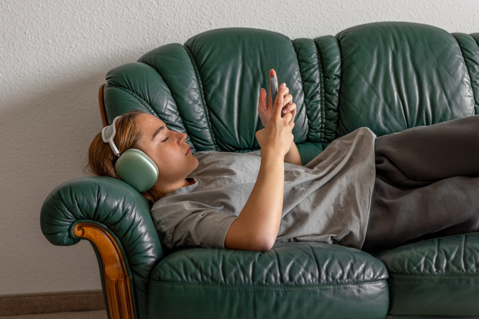 Młoda kobieta relaksująca się na zielonej kanapie w słuchawkach, przeglądająca telefon. Inspiracja na prezent dla domatora – wygoda i technologia.