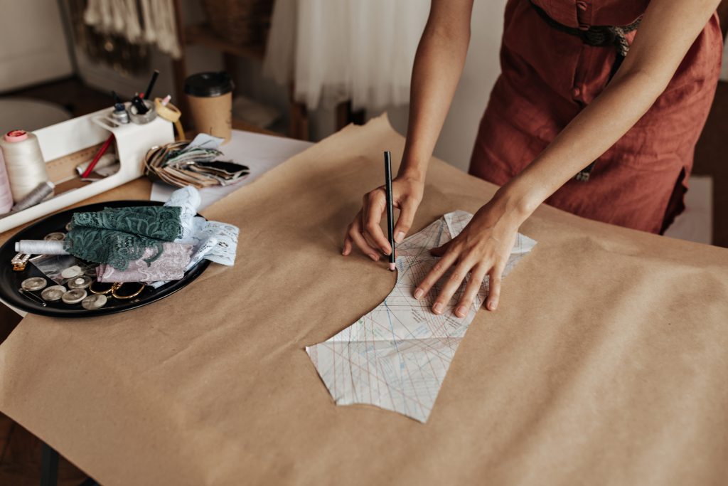 Kobieta odrysowująca wykrój krawiecki na szarym papierze w otoczeniu nici i koronek. Wyjątkowe prezenty dla fanów rękodzieła i craftu.