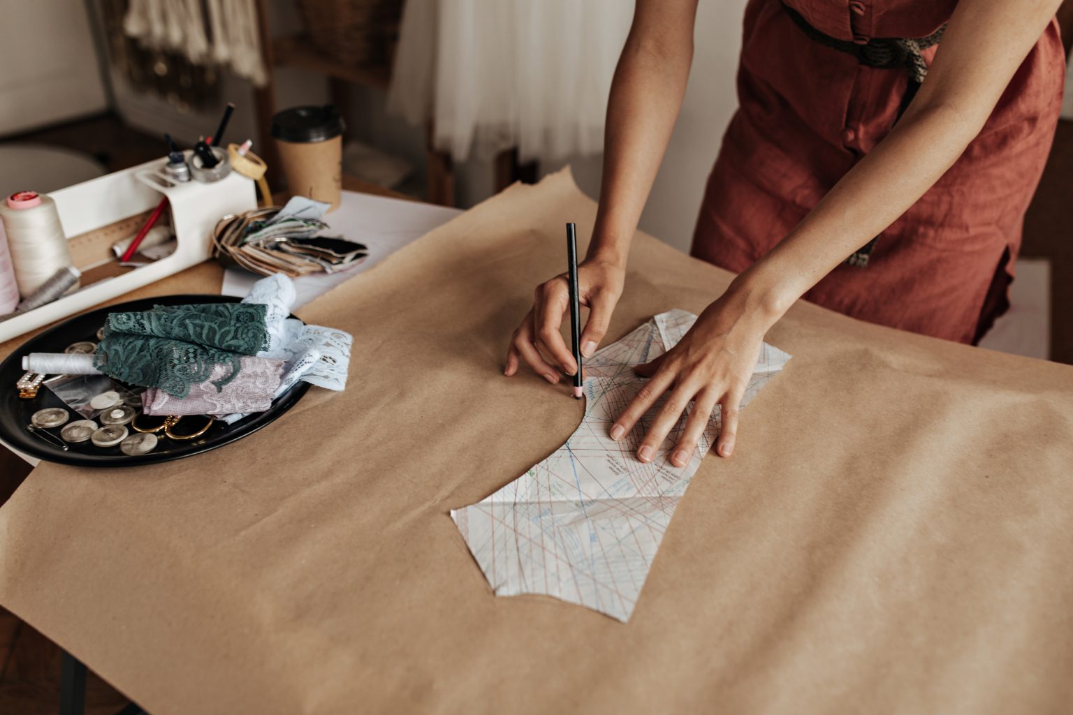 Kobieta odrysowująca wykrój krawiecki na szarym papierze w otoczeniu nici i koronek. Wyjątkowe prezenty dla fanów rękodzieła i craftu.