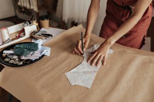 Kobieta odrysowująca wykrój krawiecki na szarym papierze w otoczeniu nici i koronek. Wyjątkowe prezenty dla fanów rękodzieła i craftu.