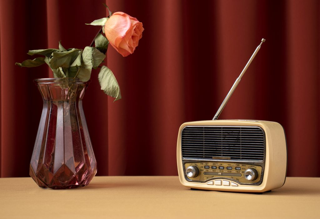 Jasnobeżowe radio w stylu retro i różowa róża w szklanym wazonie na tle bordowej zasłony. Inspiracje na prezenty w stylu vintage.