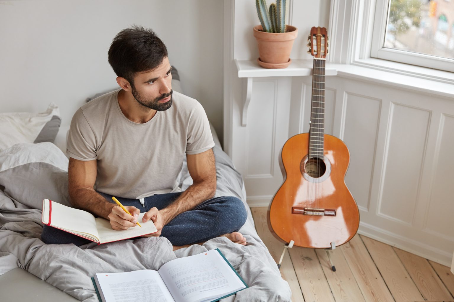 Mężczyzna siedzący na łóżku z notatnikiem i długopisem, obok niego stoi gitara klasyczna. Inspiracje na prezent na start nowego hobby