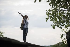 Kobieta z plecakiem stojąca na szczycie z rozłożonymi ramionami. Inspirujące prezenty zachęcające do działania i samorozwoju.